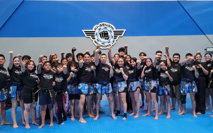 Large diverse group of adults and students in black uniforms and blue shorts posing confidently in a martial arts studio.